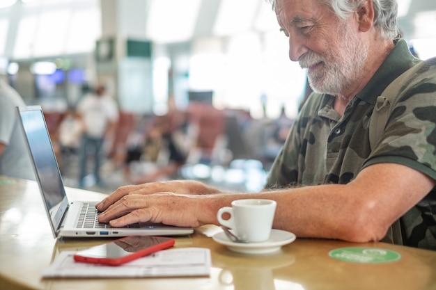 Hombre viajero en el aeropuerto esperando trabajos de embarque en una computadora portátil, trabajo remoto de blogger senior, una taza de café y un teléfono móvil sobre la mesa
