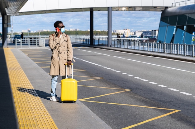 Hombre viajero afro se encuentra en la terminal del aeropuerto, llamando al móvil y esperando un taxi, use mascarilla.