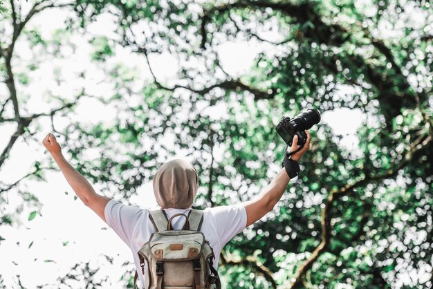 Hombre viajero asiático con mochila sosteniendo una cámara y haciendo un gesto feliz en el bosque con espacio de copia Fotógrafo de viajes Éxito vocacional y concepto de vacaciones