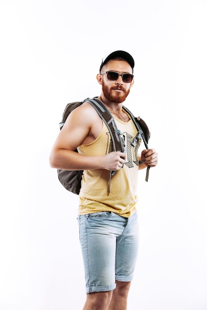 Hombre viajero barbudo con mochila en una gorra y gafas de sol mirando a la cámara sobre fondo blanco.