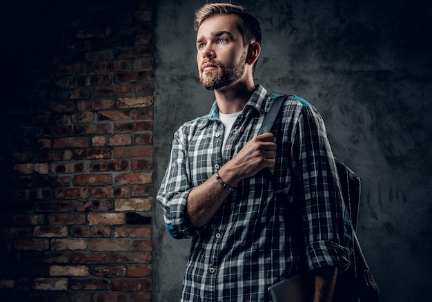 Un hombre viajero barbudo vestido con una camisa de lana con mochila posando sobre fondo gris.