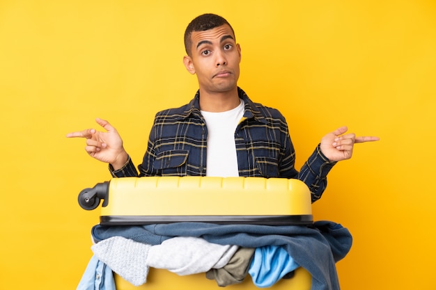 Hombre viajero con una maleta llena de ropa sobre pared amarilla aislada apuntando a los laterales que tienen dudas