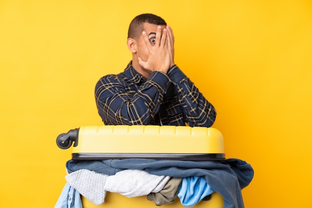 Hombre viajero con una maleta llena de ropa sobre la pared amarilla aislada que cubre los ojos y mirando a través de los dedos