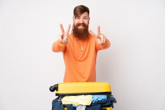 Hombre viajero con una maleta llena de ropa sobre pared blanca sonriendo y mostrando el signo de la victoria