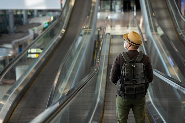 El hombre viajero con mascarilla protege el coronavirus de la gripe. Turista en la terminal del aeropuerto de pie en la escalera mecánica. Durante la pandemia debe haber autocuarentena de distanciamiento social.