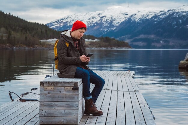 Hombre viajero mirando al teléfono sentado en el muelle de madera un fondo de una montaña y un lago