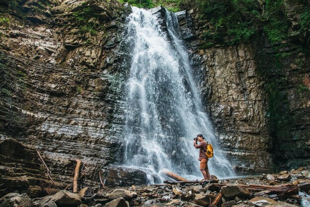 Hombre viajero con una mochila amarilla de pie sobre el fondo de una cascada hace un paisaje fotográfico