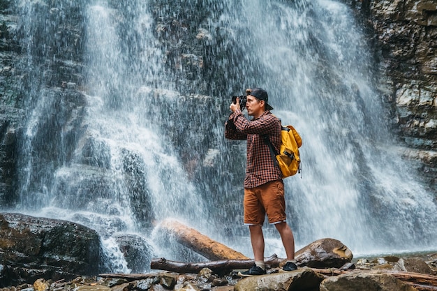 Hombre viajero con una mochila amarilla de pie sobre el fondo de una cascada hace un paisaje fotográfico