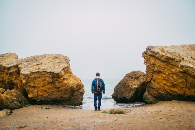 Hombre viajero con mochila de pie en la playa de arena en medio de las rocas contra el fondo del mar