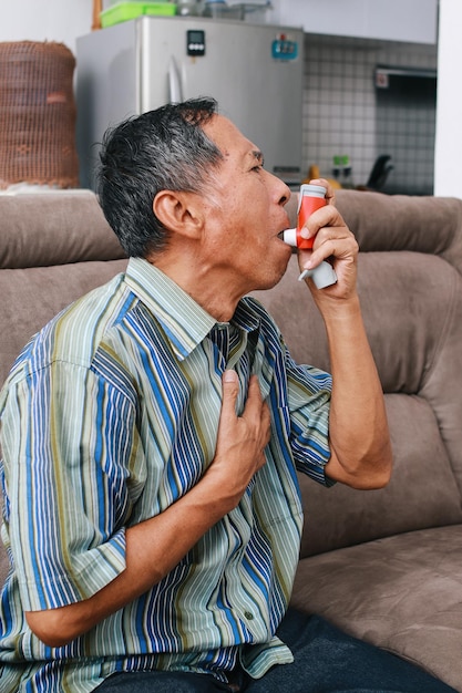 Foto hombre viejo sentado en el sofá sosteniendo el pecho con dificultades para respirar y usando inhalador de asma