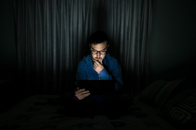 Hombre viendo series de televisión en una tableta sentada en una cama en la noche en casa