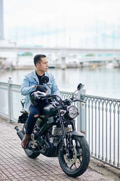 Hombre vietnamita serio seguro sentado en una motocicleta en el puente y mirando el agua