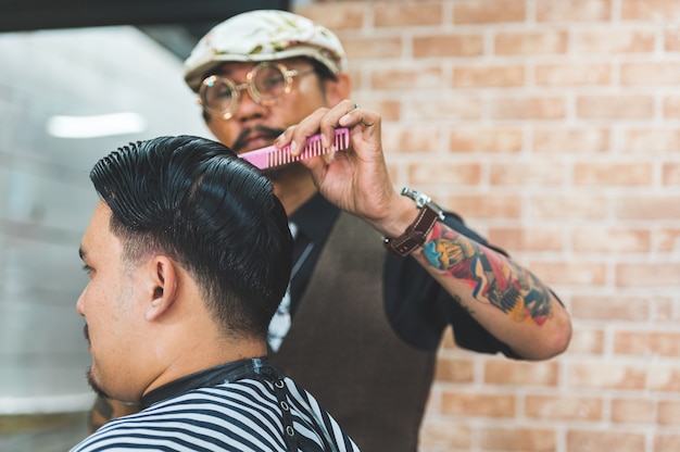 Los hombres adultos estilista elegante con tatuajes y bigote cortando el cabello del cliente en peluquería