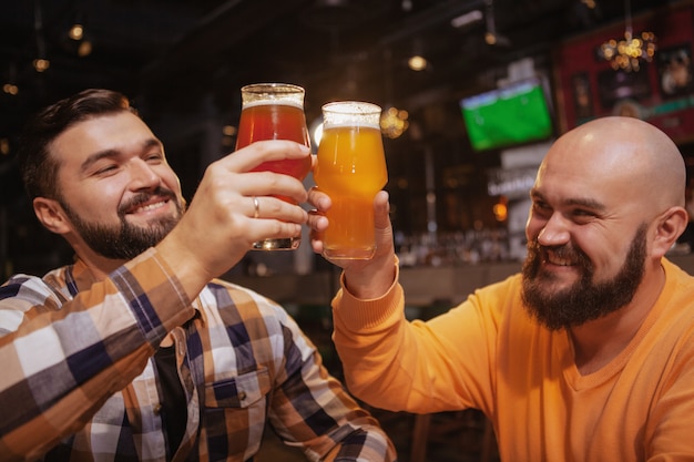 Foto hombres barbudos guapos tintineando vasos de cerveza, celebrando en el pub