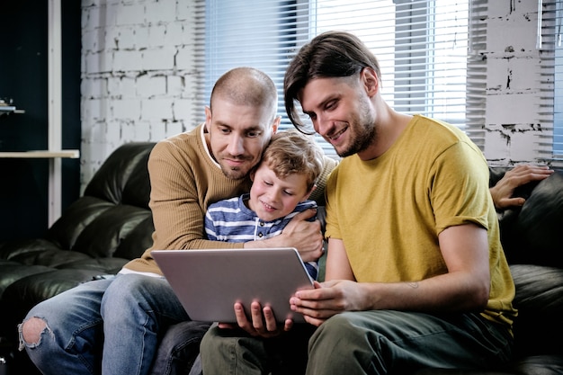 Hombres cariñosos y niño lindo viendo videos en línea en la sala de estar