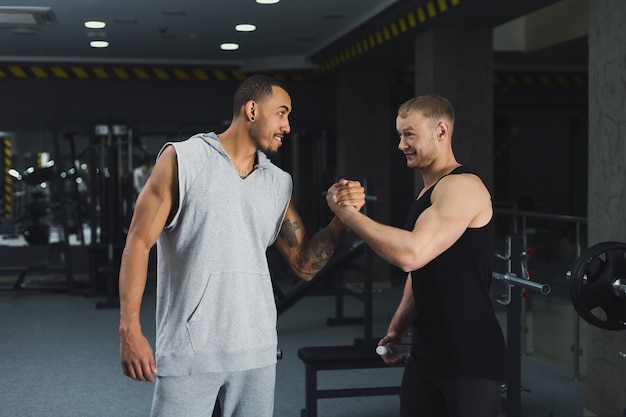 Hombres confiados musculosos que sacuden las manos. Exitosos instructores de fitness multiétnicos en el gimnasio, cartel de motivación deportiva, espacio de copia