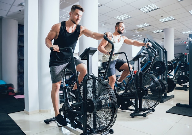 Hombres jóvenes con cuerpo musculoso usando bicicleta de aire para entrenamiento cardiovascular en el gimnasio de entrenamiento cruzado.