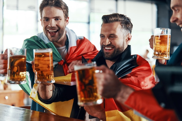 Hombres jóvenes felices cubiertos de banderas internacionales brindando con cerveza mientras ven el juego deportivo en el pub