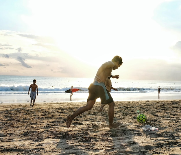 Hombres jóvenes jugando al fútbol en la arena en la playa contra el cielo durante la puesta de sol