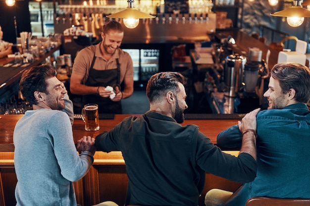 Hombres jóvenes sonrientes en ropa casual bebiendo cerveza mientras están sentados juntos en el pub