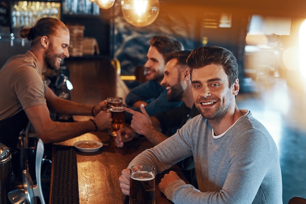 Hombres jóvenes sonrientes en ropa casual bebiendo cerveza y uniéndose juntos mientras están sentados en el pub