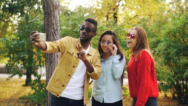 Hombres y mujeres jóvenes con estilo se toman selfie con gafas de sol posando y sonriendo sosteniendo el teléfono inteligente durante la caminata en el parque Concepto de personas y tecnología