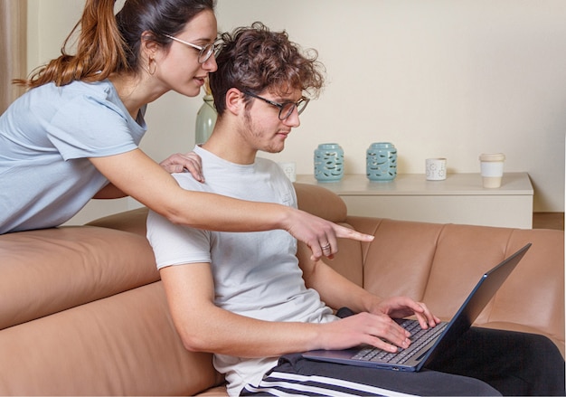 Hombres y mujeres jóvenes que trabajan con la computadora portátil mientras están sentados en el sofá beige en casa. Las mujeres jóvenes señalan la computadora portátil con la mano.