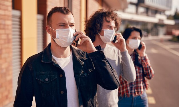 Hombres y mujeres modernos con máscaras médicas hablando por teléfonos inteligentes y mirando hacia otro lado mientras están parados en la calle durante la pandemia de coronavirus