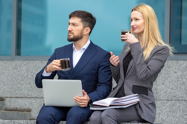 hombres y mujeres de negocios sentados en las escaleras de un edificio de negocios con documentos y una laptop en sus manos tomando café y discutiendo el plan de trabajo de negocios