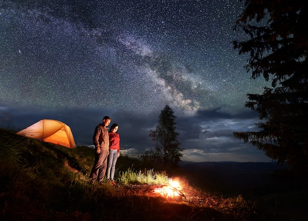 Hombres y mujeres turistas están de pie junto al campamento y la carpa naranja, mirando la hoguera bajo el hermoso cielo estrellado.