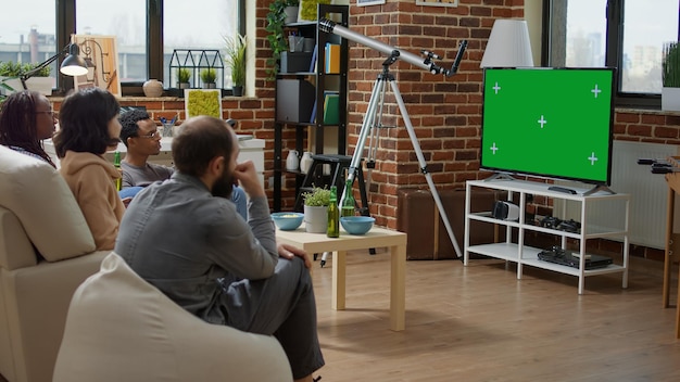 Hombres y mujeres viendo televisión con pantalla verde en la sala de estar, usando una plantilla de clave de croma aislada con espacio de copia de maqueta y fondo en blanco. Tecnología moderna en pantalla de TV. Disparo de trípode.