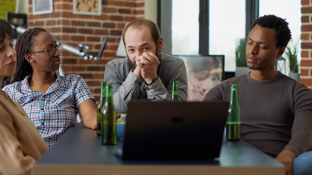 Hombres y mujeres viendo videos en vivo en el sitio web de las redes sociales, usando tecnología portátil para ver programas de televisión juntos en la sala de estar. Gente moderna divirtiéndose con la red inalámbrica en línea.