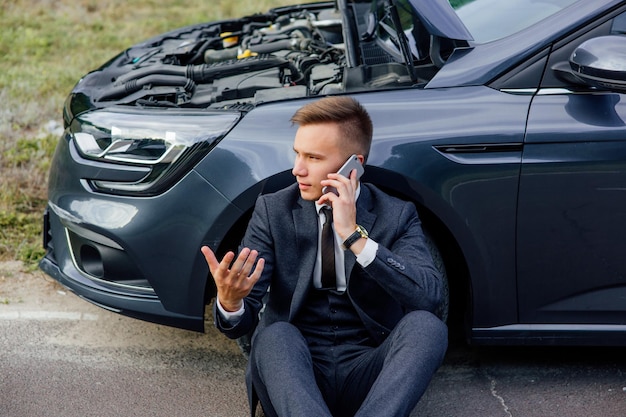 Hombres de negocios caucásicos hablando por un teléfono celular junto a un automóvil roto en una carretera rural llamando para pedir ayuda al hombre b ...