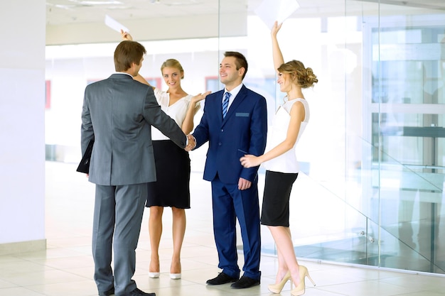 Hombres de negocios dándose la mano Dos hombres de negocios confiados dándose la mano y sonriendo mientras están de pie en la oficina junto con la gente en el fondo