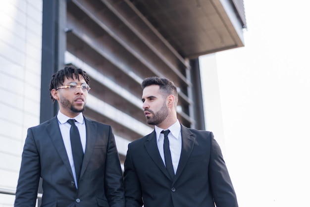 Hombres de negocios vestidos con trajes caminando y sonriendo y hablando al aire libre en un área de negocios