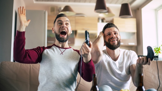 Hombres sonrientes emocionados jugando en videojuegos en la televisión en casa en el sofá. Amigos con joysticks juegan con emociones felices en las caras