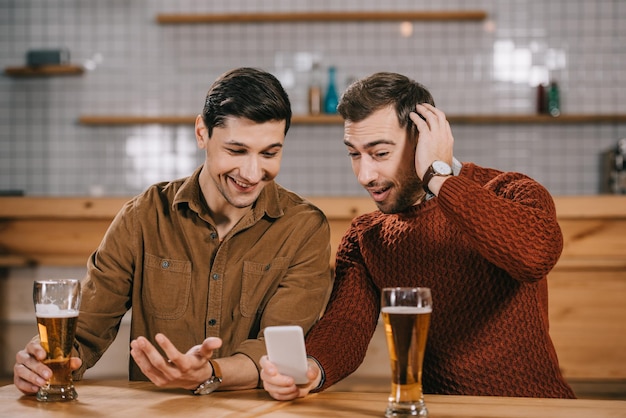 Foto hombres sorprendidos mirando el teléfono inteligente cerca de vasos con cerveza