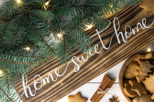 Home Sweet Home Schriftzug aus gebranntem Holz mit Lebkuchenplätzchen Christbaumzweig Zimtstangen und Anis auf weißer Oberfläche Flache Lage