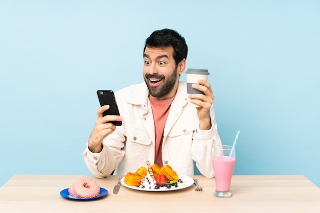 Homem à mesa tomando waffles no café da manhã e um milkshake segurando café para levar e um celular