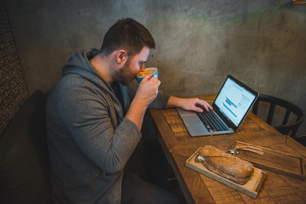 Homem a trabalhar num portátil num café, pequeno-almoço, sanduíche e café.
