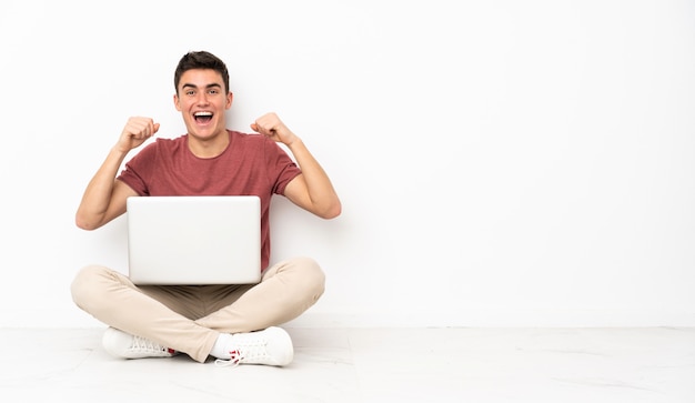 Homem adolescente sentado na flor com seu laptop comemorando uma vitória na posição de vencedor