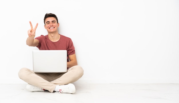 Homem adolescente sentado na flor com seu laptop sorrindo e mostrando sinal de vitória