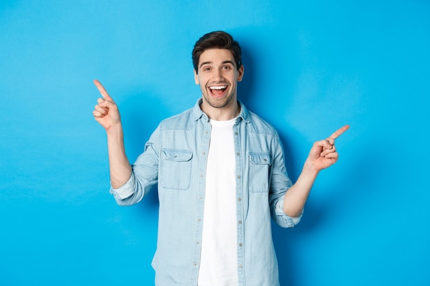 Homem adulto alegre sorrindo, apontando o dedo para o lado, mostrando faixas promocionais à esquerda e à direita, encostado na parede azul