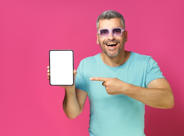 Homem adulto animado, 30 e 40 anos, segura um enorme tablet digital vestindo camisa azul casual e óculos de sol isolados em fundo rosa Homem com telefone, foto de estúdio, anúncio de aplicativo móvel