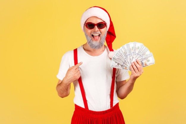 Homem adulto animado extremamente feliz com chapéu de papai noel e óculos de sol elegantes, apontando o dedo para dólares na mão, vantagens e bônus nos feriados. Tiro de estúdio interior isolado em fundo amarelo