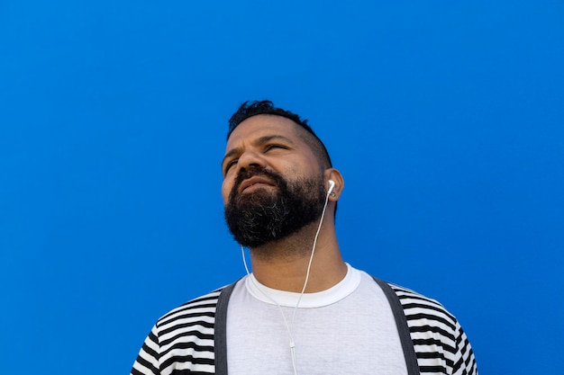 Homem adulto 39 anos com barba média curta lisa Ouvi música usando fones de ouvido brancos Ele olha para cima do lado direito Vestido com suéter listrado camiseta branca fundo de parede azul Espaço para copiar