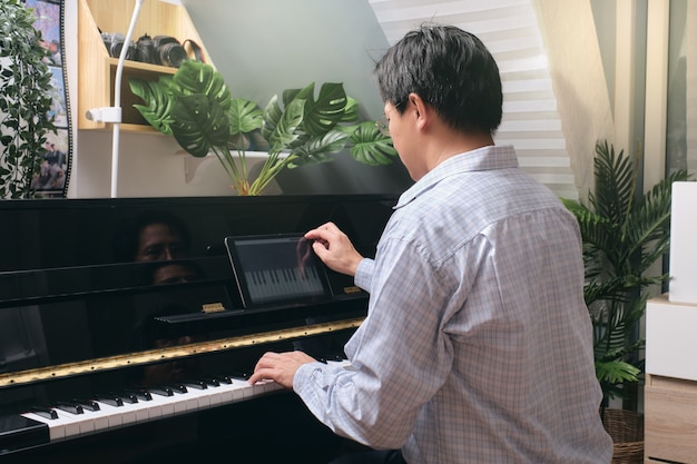 Homem adulto aprendendo a tocar piano usando tablet digital com aula on-line e curso na sala de estar em casa. feliz empresário asiático relaxando tocando piano