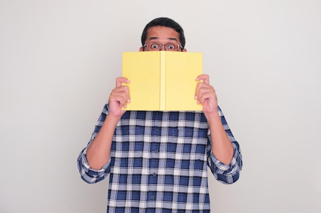 Homem adulto asiático espreitando por trás de um livro com expressão de olhos chocados