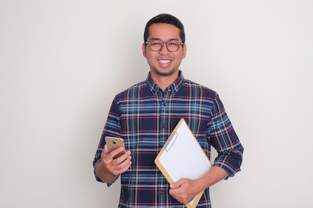 Foto homem adulto asiático sorrindo feliz enquanto segura celular e papel no clipboard