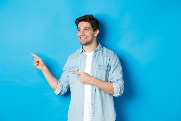 Homem adulto bonito apresenta o produto, olhando e apontando o dedo para a esquerda, promovendo algo contra a parede azul